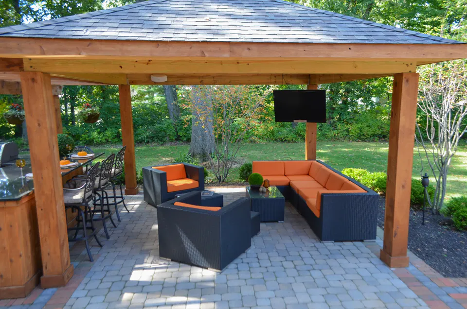 Covered pavilion interior with television wall, orange lounge seating, paver floor, wood posts, and backyard view.